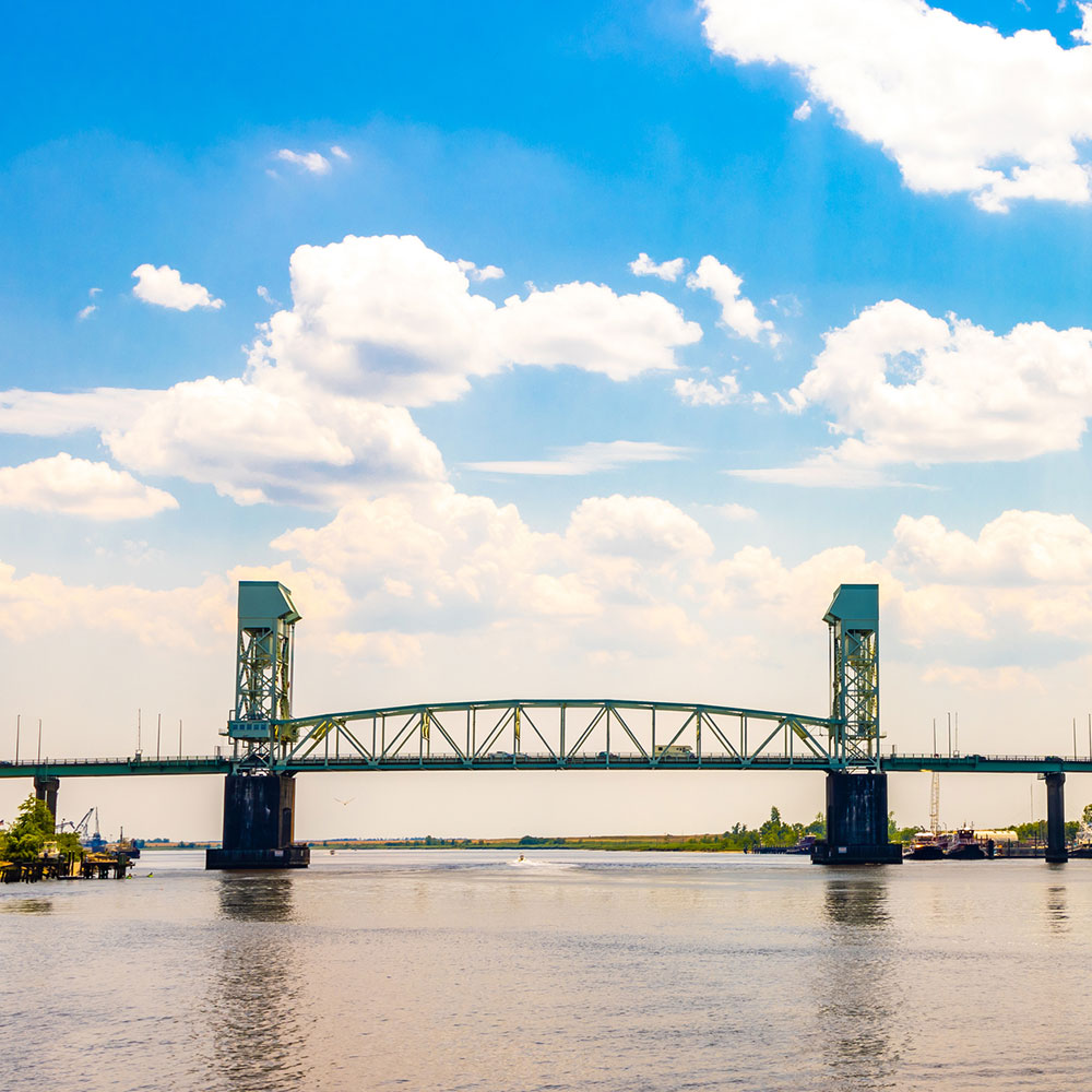 The Cape Fear Memorial Bridge, an iconic arch bridge crossing the Cape Fear River in Wilmington, NC, during a partly cloudy day.
