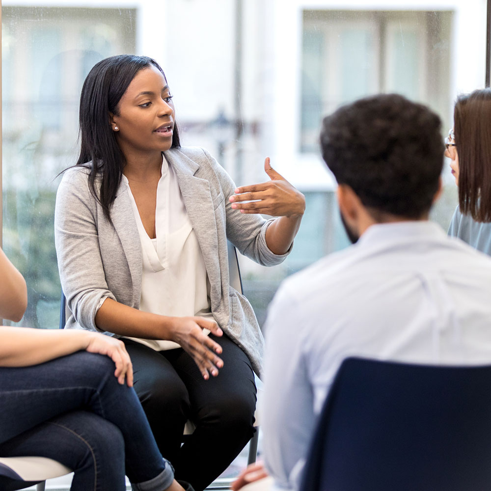 A young female counselor gestures as she sits in a circle with clients during a group therapy session and speaks. She is looking at the female client next to her.